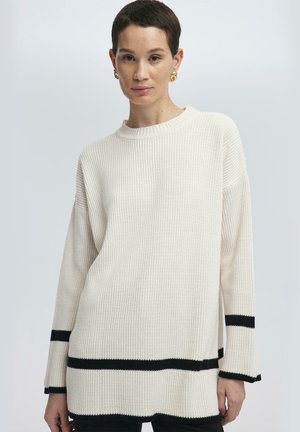Vit ribbad stickad tröja med långa ärmar, svarta ränder vid fållen och muddarna, samt rund halsringning, som visar en texturerad yta.