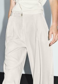 Pantalons larges beige clair en tissu doux, avec une taille haute, des plis et un unique bouton décoratif à l'avant.