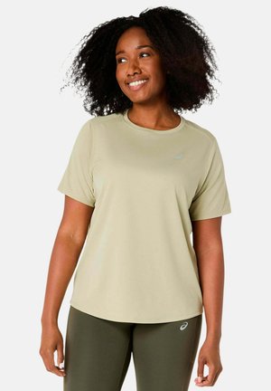 Lächelnde Frau mit lockigem Haar, die ein hellbeiges Kurzarmshirt und olivgrüne Leggings trägt und vor einem schlichten Hintergrund steht.