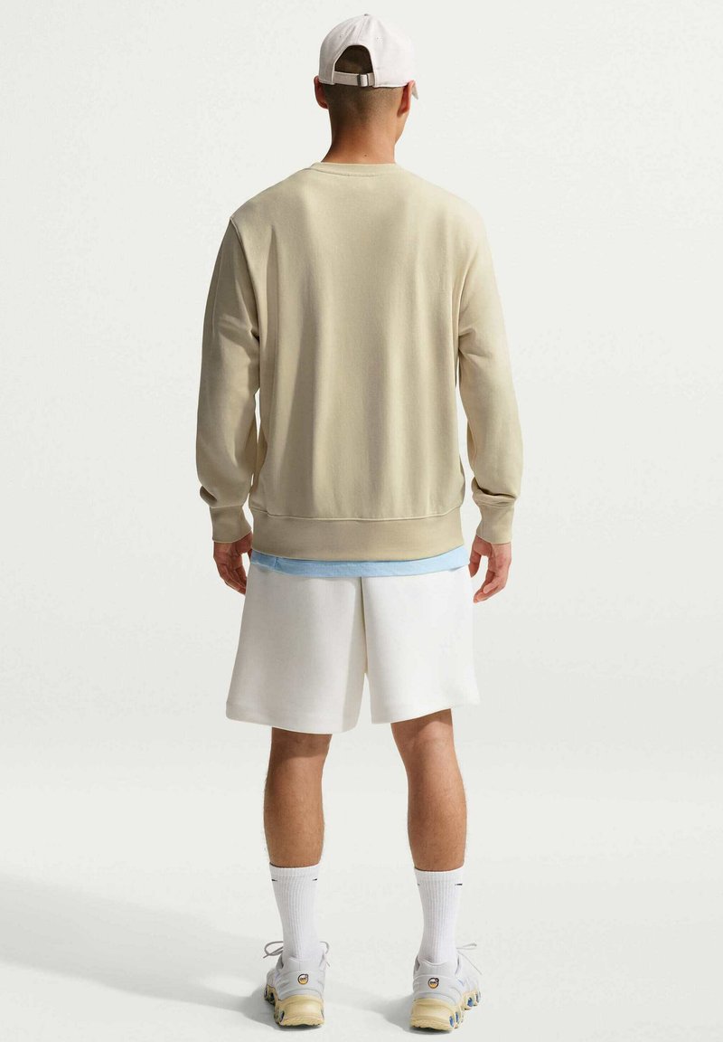 Sweat-shirt beige à manches longues, avec poignets et ourlet côtelés, porté sur une couche bleu clair, associé à un short blanc et des baskets blanches.