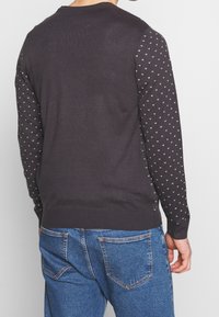 Maglione grigio scuro con maniche a fantasia caratterizzate da piccoli cuori bianchi. Polsini e orlo a costine, design aderente. Indossato sopra jeans di denim blu.