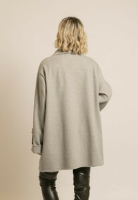 Manteau en laine oversize gris avec un col, doté de manches longues et d'une coupe ample, associé à un pantalon en cuir noir. Pas de fermetures visibles.