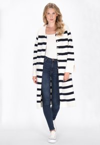 Cardigan lungo a righe in crema e blu navy, con una texture a coste, chiusura con bottoni e maniche ampie, abbinato a jeans aderenti e sneakers bianche.