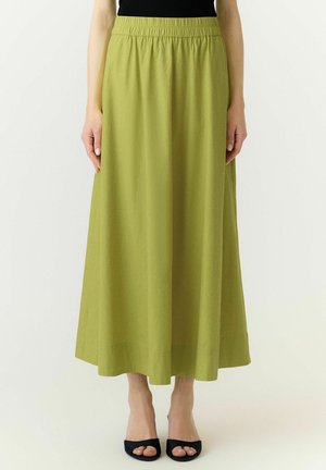 Femme portant une longue jupe vert olive avec une taille élastique et des talons noirs ouverts, debout devant un fond uni.