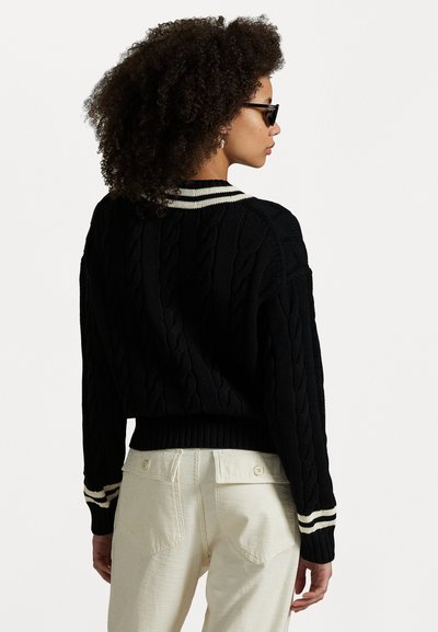 Pull en maille noire avec col à rayures. Conception courte, ourlet et poignets côtelés. Porté sur un pantalon crème.