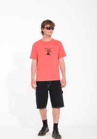 T-shirt en coton corail avec un design graphique noir, associé à un short cargo noir, des chaussettes noires et des chaussures noires. Modèle portant des lunettes de soleil.