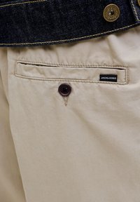 Μπεζ βαμβακερό παντελόνι με πίσω τσέπη που διαθέτει σκοτεινό κουμπί. Μια μαύρη ετικέτα με λευκό κείμενο εμφανίζει το "JACK & JONES".