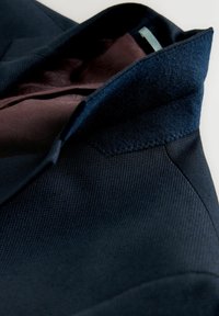 Marineblauer strukturierter Blazer mit einem glatten, braun gemusterten Innenfutter. Der Kragen besteht aus einem filzähnlichen Material mit markanten Nähten.