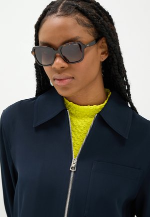 Persona con capelli intrecciati che indossa grandi occhiali da sole scuri, una giacca blu navy con zip e un top giallo brillante e strutturato sotto.