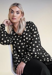 Svart blus med vita prickar, v-ringning och elastiska puffärmar. Slät tyg med avslappnad passform, parat med svarta byxor.