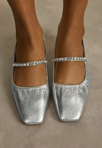 Hopeisia ballerina-kenkiä, joissa on sileä metallinen pinta, nelikulmainen kärki ja koristeltu kimaltavalla strassihihnalla jalkapöydän kohdalla.