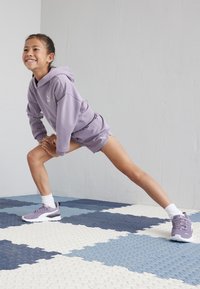 Conjunto de sudadera y pantalones cortos lilas con el logotipo blanco de Puma. Modelo con zapatillas lilas a juego y calcetines blancos sobre un suelo texturizado y con patrones.
