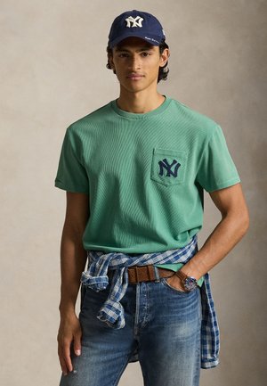 Grønn bomulls-t-skjorte med en lomme foran og mørkeblått NY-logo, kombinert med blå denimjeans og en rutete skjorte knyttet rundt livet. Mørkeblå caps brukes.