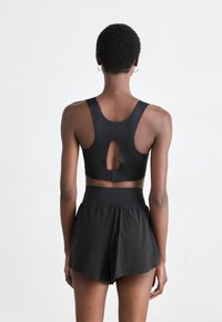 Persona con cabello corto y rizado que lleva puestos unos pantalones cortos negros de cintura alta y un top corto negro de tirantes tipo racerback con un corte en forma de ojo de cerradura en la espalda.