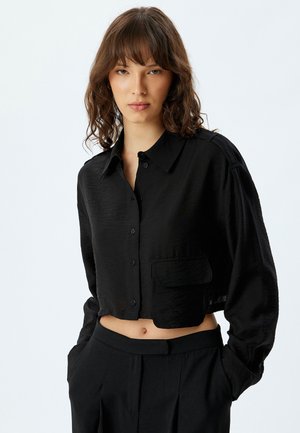 Zwarte, korte cropped shirt van lichtgewicht, textuurstof. Heeft een knoopsluiting aan de voorkant, een kraag en een enkele borstzak. Lange mouwen met manchetten.
