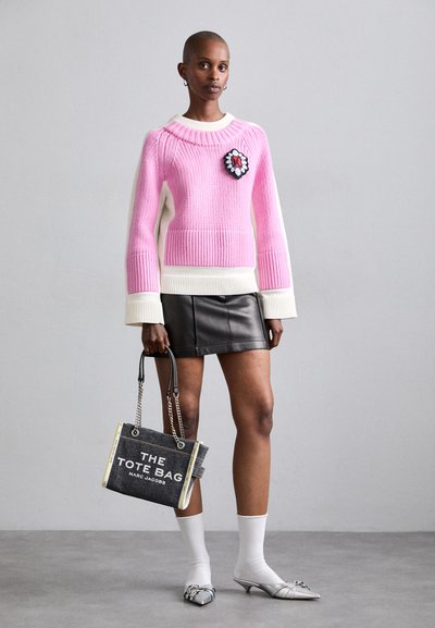 Pull rose tricoté avec des accents blancs, un appliqué noir et rouge, associé à une jupe en cuir noir et un sac fourre-tout gris avec du texte blanc.