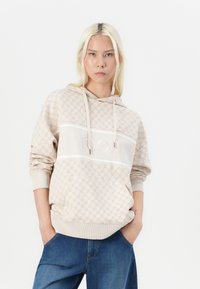 Beige Kapuzenpullover mit strukturiertem Muster, ausgestattet mit einem zentralen weißen Streifen und einem geprägten Logo. Rippbündchen und Saum, kombiniert mit blauen Jeanshosen.