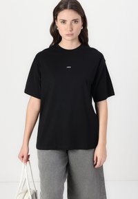 Black cotton t-shirt with short sleeves and round neckline, featuring a small white logo on the chest. Gray pants and white bag visible.