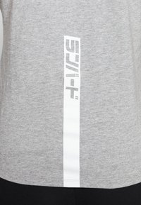 Camiseta gris de manga corta con una raya vertical blanca que presenta un logo impreso. Tela suave y texturizada; diseño clásico de cuello redondo.