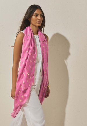 Woman wearing sleeveless white outfit and bright pink scarf with gold floral pattern, walking against a plain beige wall.
