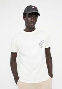 T-shirt en coton blanc à manches courtes et col rond, arborant un petit logo noir sur la poitrine gauche. Casquette en denim avec des accents de logo.