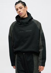 Fekete és szürke Nike Tech túlnyomó kapucnis pulóver magas gallérral, puha textúrával, diszkrét logóval és egyedi bolyhos panel díszítéssel az ujjakon.