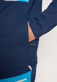 Sweatshirt bleu marine texturé avec une poche latérale et un motif en bleu clair contrastant. Il présente des poignets côtelés et un ourlet arrondi.