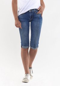 Capris de mezclilla con corte ajustado, puños enrollados y un lavado azul medio. Presentan bolsillos estándar y cremallera. Combinados con zapatillas blancas.