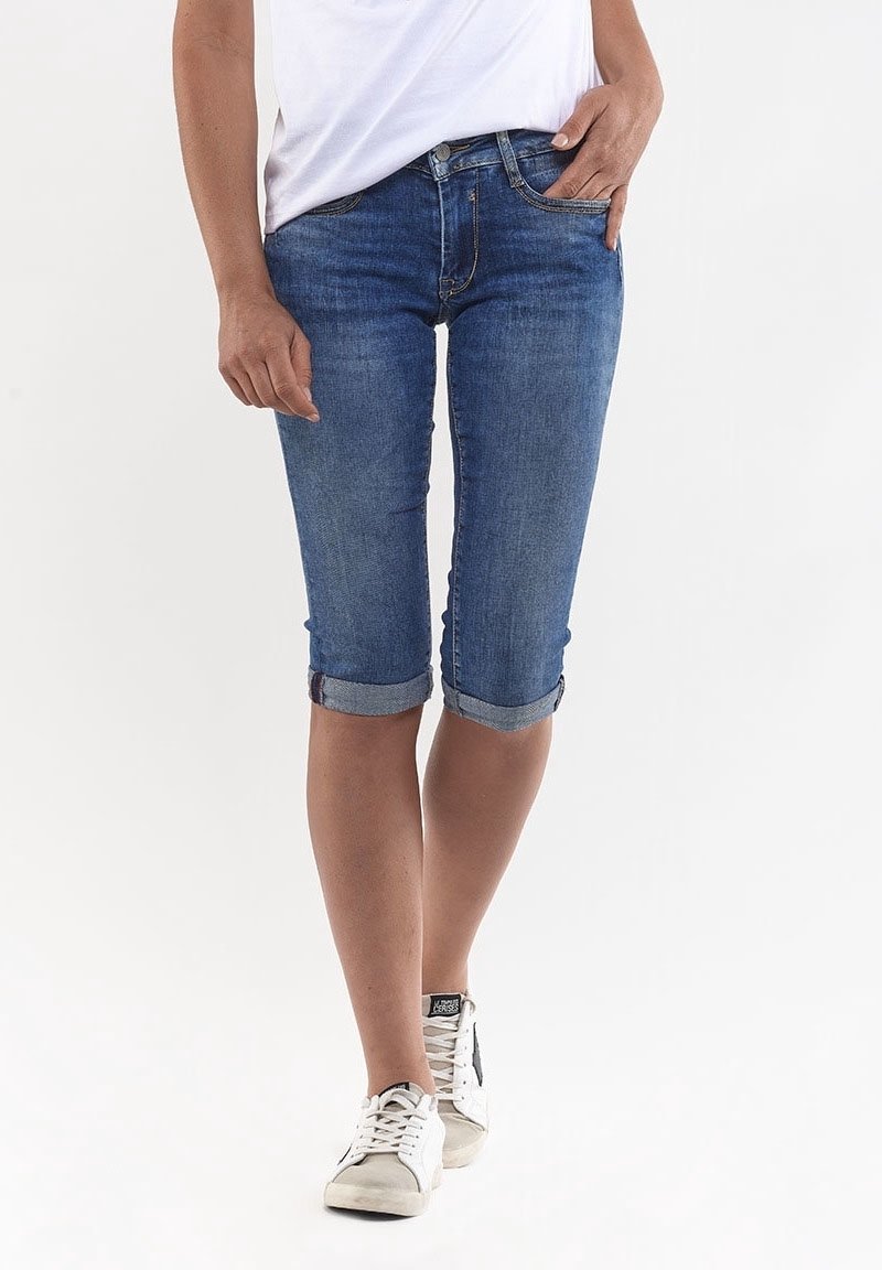 Capris de mezclilla con corte ajustado, puños enrollados y un lavado azul medio. Presentan bolsillos estándar y cremallera. Combinados con zapatillas blancas.