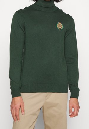 Grüner Rollkragenpullover aus weichem Strickstoff. Mit einem goldenen und grünen gestickten Emblem auf der Brust. Mit gerippten Bündchen und Saum.