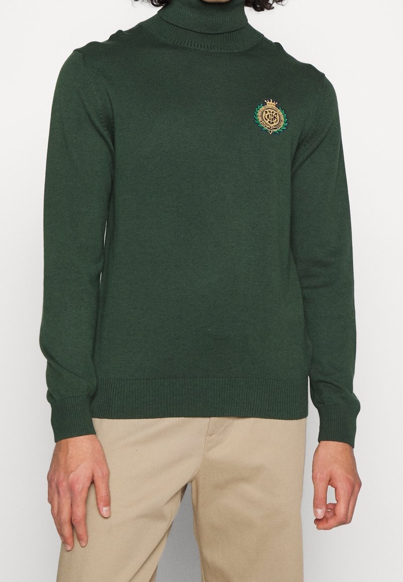Grüner Rollkragenpullover aus weichem Strickstoff. Mit einem goldenen und grünen gestickten Emblem auf der Brust. Mit gerippten Bündchen und Saum.