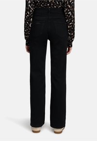 Persoon in zwarte hoge taille spijkerbroek met rechte pijpen en een lang mouw leopardprint shirt, gezien vanaf de achterkant tegen een effen achtergrond.