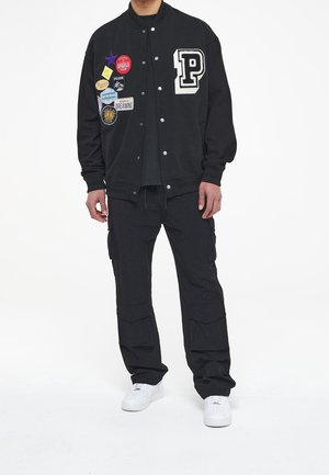 Hombre con chaqueta varsity negra con parches coloridos, pantalones cargo negros y zapatillas blancas, de pie contra un fondo blanco.