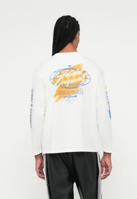 Vyras su pintomis plaukais, vilkintis baltą ilgų rankovių Adidas Originals marškinėlį su mėlynomis ir oranžinėmis grafikomis, juodas Adidas kelnes su baltais dryžiais.