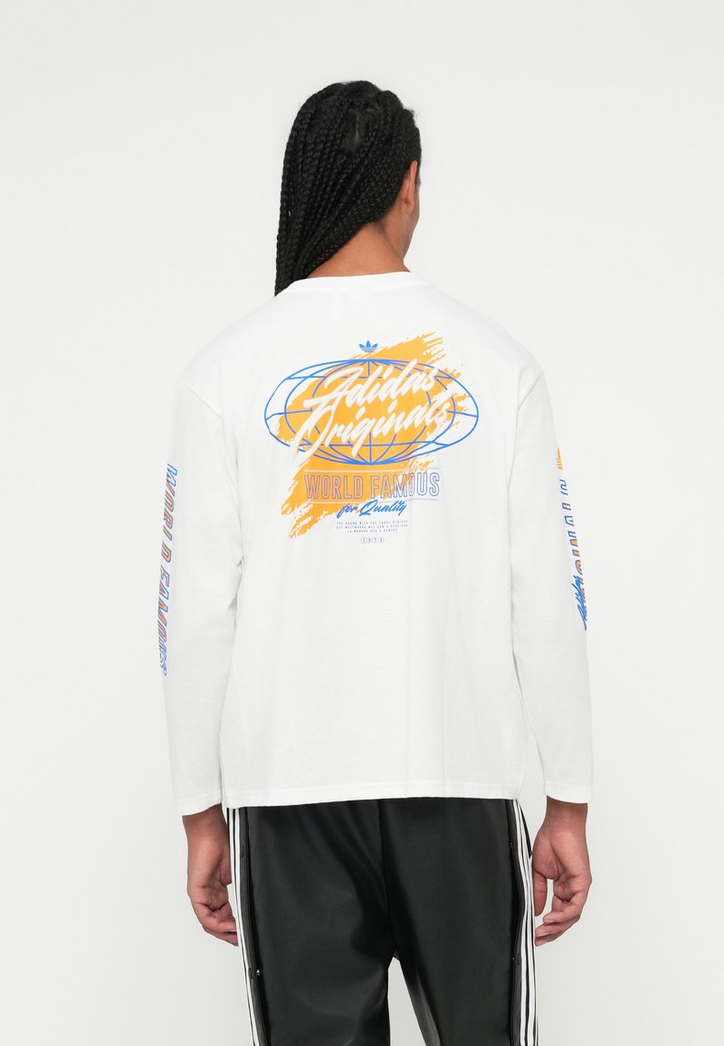 Vyras su pintomis plaukais, vilkintis baltą ilgų rankovių Adidas Originals marškinėlį su mėlynomis ir oranžinėmis grafikomis, juodas Adidas kelnes su baltais dryžiais.