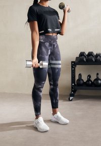 Top de entrenamiento negro recortado con el texto "MÁS FUERTES", leggings gris tie-dye con rayas horizontales y zapatillas deportivas blancas sosteniendo mancuernas.