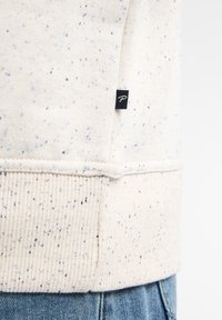 Maglione crema con macchie blu, orlo a coste e una piccola etichetta nera di lato. Tessuto morbido con una vestibilità rilassata.