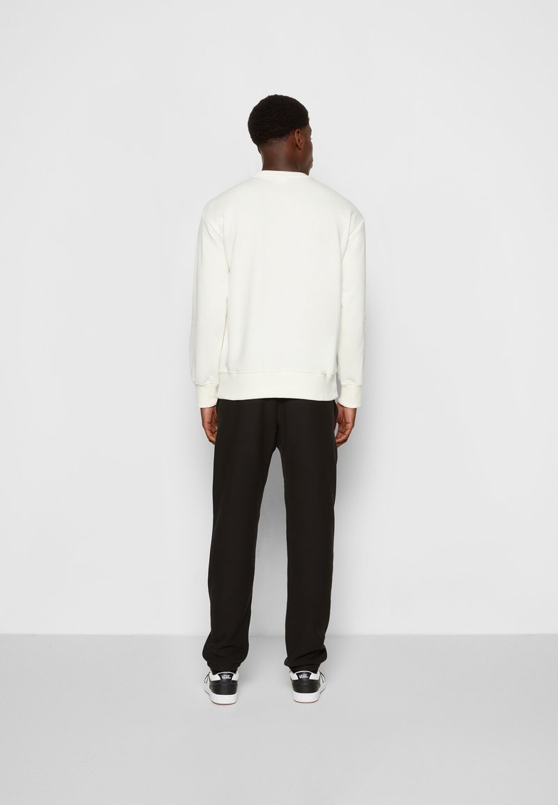 Felpa bianca con polsini a coste, abbinata a pantaloni neri. Dettaglio logo visibile sulle calzature. Sfondo semplice che mette in risalto l'abbigliamento.