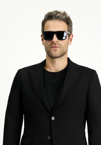 Schwarzer Blazer mit Spitzenrevers, Ein-Knopf-Verschluss und einem dezenten strukturierten Stoff, kombiniert mit schwarzen Sonnenbrillen und einem schwarzen T-Shirt.