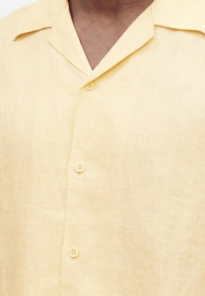Gros plan d'une personne portant une chemise en lin jaune pâle avec un col camp et trois boutons visibles fermés.