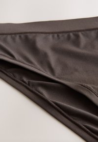 Primer plano de ropa interior tipo bikini sin costuras de color marrón oscuro con tela lisa y costuras limpias, colocada plana sobre una superficie clara.