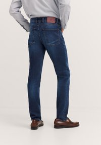 Dunkelblaue Denim-Jeans mit geradem Schnitt, Gesäßtaschen und einem Leder-Label-Patch am Bund. Kombiniert mit braunen Schuhen.