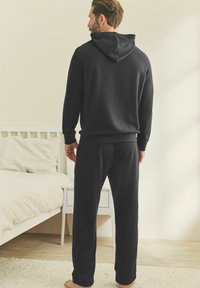 Donkergrijze hoodie en joggingset van textuurstof, met een capuchon en elastische manchetten, van de achterkant weergegeven tegen een neutrale achtergrond.