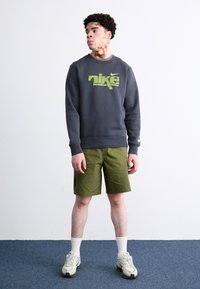 Γκρι φούτερ με νεον πράσινο λογότυπο "Nike", ελαιόχρωμες βερμούδες, λευκές κάλτσες και ανοιχτόχρωμα αθλητικά παπούτσια. Το μοντέλο στέκεται σε ένα μπλε χαλί.