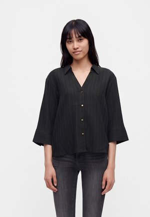 Jeune femme aux cheveux noirs et raides, portant une chemise noire ample boutonnée et un jean gris foncé, debout devant un fond blanc uni.