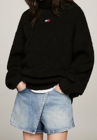 Person i svart chunky stickad tröja med Tommy Jeans-logotyp och ljusblå denimkortbyxor med händerna i fickorna.