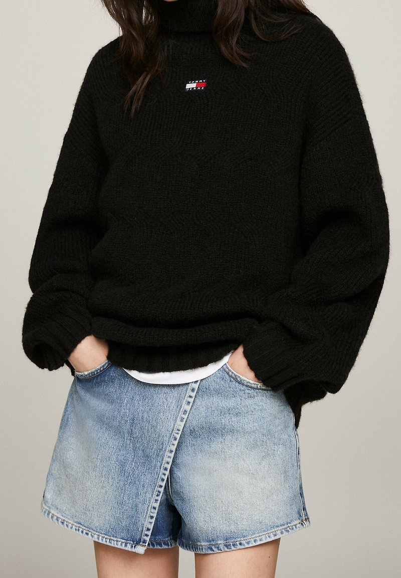 Person i svart chunky stickad tröja med Tommy Jeans-logotyp och ljusblå denimkortbyxor med händerna i fickorna.