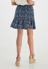 Falda floral en navy con patrones azules y verdes, que presenta un dobladillo con volantes y una cintura elástica; combinada con sandalias de cuña en tono beige.