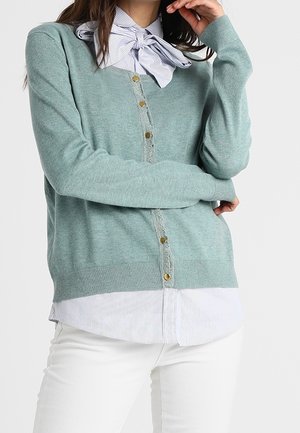 Kvinde iført en lysegrøn cardigan med knapper over en stribet skjorte med stor sløjfe ved kraven, kombineret med hvide bukser, poserer med armene krydset.