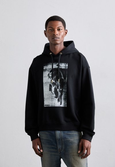 Jeune homme portant un sweat à capuche noir avec un imprimé de sculpture classique en niveaux de gris et un jean bleu, se tenant devant un fond gris clair uniforme.